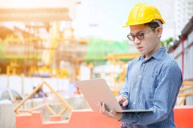 Genç Asyalı mühendisler inşaat alanında çalışıyorlar. Sarı kask emniyeti takın. Elle tutulan bilgisayar dizüstü