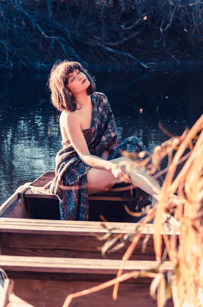 Young sexy woman on boat at sunset. The girl has a flower wreath on her head, relaxing and sailing on river. Beautiful body and face. Fantasy art photography. Concept of female beauty, rest in the