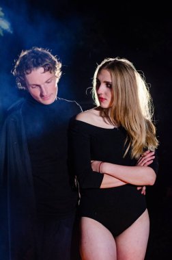 Siyah elbiseli genç seksi bir kadın ve dumanlı bir arka planda poz veren bir adam.