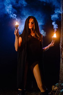 Gizemli genç kız siyah pelerin giyiyor, cadıların Şenlik ateşinin yanındaki ormanda Şabat günü
