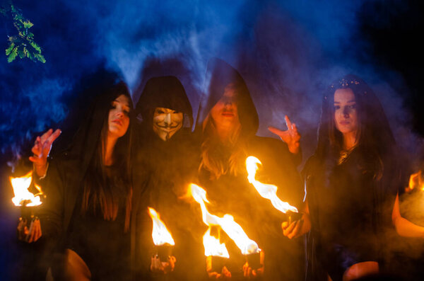загадочные молодые девушки и мужчина в черных плащах, шабаш ведьмы в лесу ночью у костра