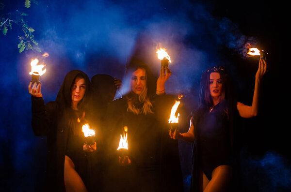 mystery young girls and man wearing black cloaks, witch's sabbath in the woods at the night by the bonfire