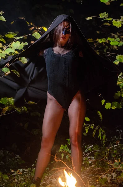 一个身穿黑色斗篷的神秘少女 一个巫婆的安息日在篝火边的树林里 — 图库照片