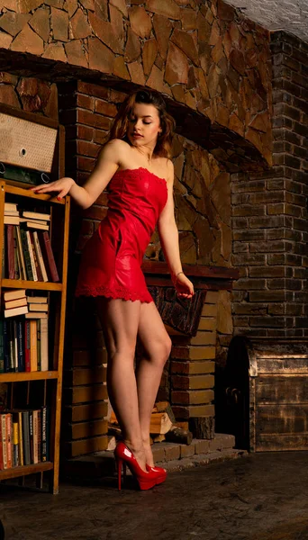 Чудові сексуальні жінки в довгій червоній вечірній сукні, що сидить у кріслі біля декоративного місця вогню. Красива новорічна та різдвяна сцена, портрет модної моделі . — стокове фото