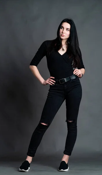 Retrato de una joven morenita sentada en una silla de madera con piernas cruzadas. Elegante y elegante chica con pantalones de pierna ancha. Estudio de rodaje sobre fondo gris —  Fotos de Stock