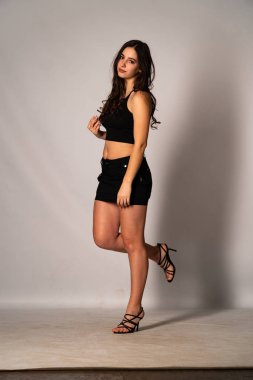 Siyah elbiseli, siyah saçlı, açık renk stüdyo arka planında poz veren güzel bir kadın. 