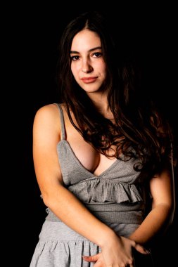 Koyu kahverengi saçlı, gri elbiseli, esmer, güzel bir kadın. Stüdyo arka planında poz veriyor.  