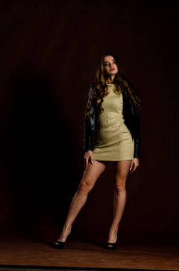 Deri ceketli, parlak elbiseli, siyah arka planda yüksek topuklu, çekici bir kadın. 