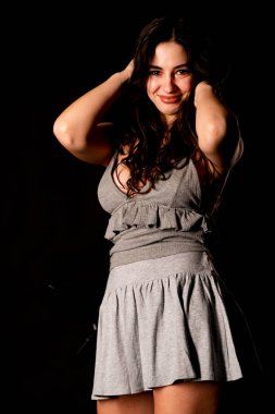 Koyu kahverengi saçlı, gri elbiseli, esmer, güzel bir kadın. Stüdyo arka planında poz veriyor.  