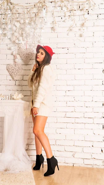 Kırmızı şapkalı ve kalın kazaklı güzel seksi kadın tuğla duvarda poz veriyor. 
