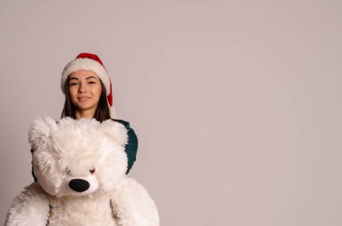 Küçük bir arka planda oyuncak ayıyla Noel Baba şapkalı genç bir kız.