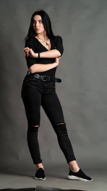 Yüksek tahta koltukta bacak bacak üstüne atmış esmer bir kadının portresi. Geniş paçalı pantolon giyen şık bir kız. Gri arkaplanda stüdyo çekimi