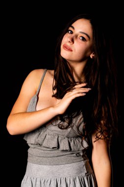 Koyu kahverengi saçlı, gri elbiseli, esmer, güzel bir kadın. Stüdyo arka planında poz veriyor.  