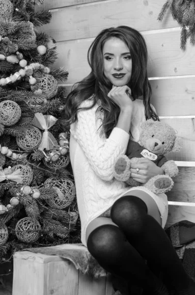 Hermosa mujer sexy con árbol de Navidad en el fondo sentado en una silla elegante en un paisaje acogedor. Retrato de niña posando vestido blanco ajustado bastante corto. Atractiva morena femenina, tiro de interior . — Foto de Stock
