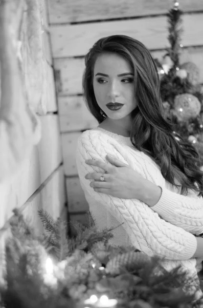Beautiful sexy woman with Xmas tree in background sitting on elegant chair in cozy scenery. Portrait of girl posing pretty  short tight fit white dress. Attractive brunette female, indoor shot. — Stok fotoğraf