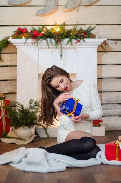 Schöne sexy Frau mit Weihnachtsbaum im Hintergrund sitzt auf eleganten Stuhl in gemütlicher Landschaft. Porträt von Mädchen posiert ziemlich kurz eng anliegendes weißes Kleid. attraktive brünette weiblich, indoor shot. — Stockfoto