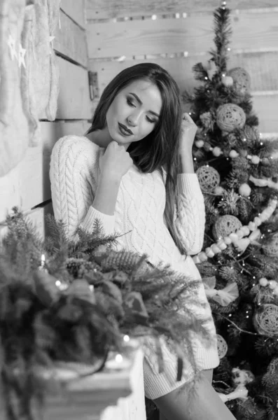 Beautiful sexy woman with Xmas tree in background sitting on elegant chair in cozy scenery. Portrait of girl posing pretty  short tight fit white dress. Attractive brunette female, indoor shot. — Stok fotoğraf