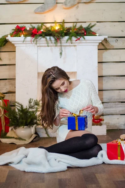 Piękna seksowna kobieta z choinką w tle siedzi na eleganckim krześle w przytulnej scenerii. Portret dziewczyny pozującej dość krótką, ciasno dopasowaną białą sukienkę. Atrakcyjne brunetka kobieta, wewnątrz strzał. — Zdjęcie stockowe