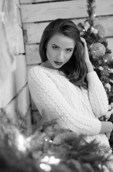 Beautiful sexy woman with Xmas tree in background sitting on elegant chair in cozy scenery. Portrait of girl posing pretty  short tight fit white dress. Attractive brunette female, indoor shot. — Stok fotoğraf