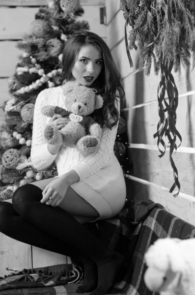 Beautiful sexy woman with Xmas tree in background sitting on elegant chair in cozy scenery. Portrait of girl posing pretty  short tight fit white dress. Attractive brunette female, indoor shot. — Stok fotoğraf