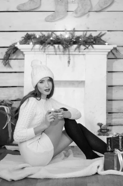 Beautiful sexy woman with Xmas tree in background sitting on elegant chair in cozy scenery. Portrait of girl posing pretty  short tight fit white dress. Attractive brunette female, indoor shot. — Stok fotoğraf