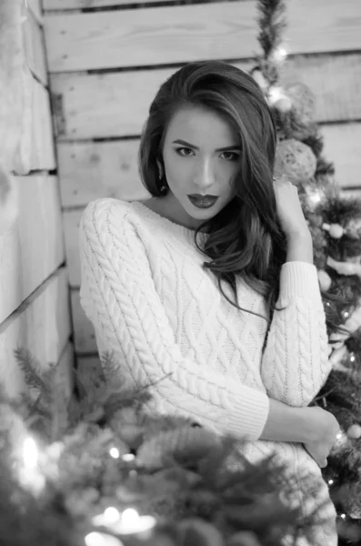 Όμορφη σέξι γυναίκα με χριστουγεννιάτικο δέντρο στο παρασκήνιο κάθεται σε κομψή καρέκλα στο ζεστό τοπίο. Πορτρέτο του κοριτσιού που θέτουν αρκετά σύντομο σφιχτό ταιριάζει λευκό φόρεμα. Ελκυστική μελαχρινή γυναίκα, εσωτερικό πλάνο. — Φωτογραφία Αρχείου