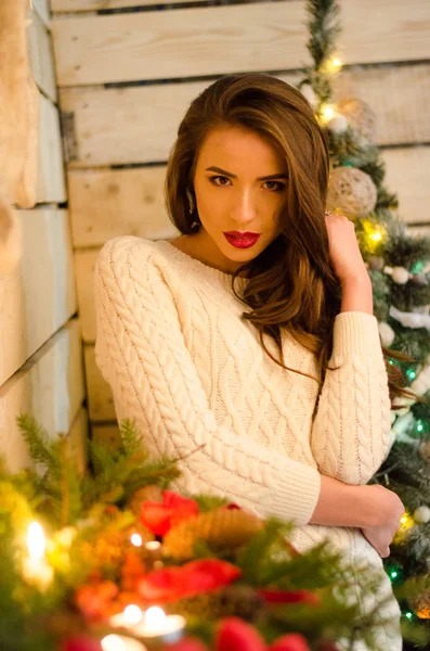 Schöne sexy Frau mit Weihnachtsbaum im Hintergrund sitzt auf eleganten Stuhl in gemütlicher Landschaft. Porträt von Mädchen posiert ziemlich kurz eng anliegendes weißes Kleid. attraktive brünette weiblich, indoor shot. — Stockfoto