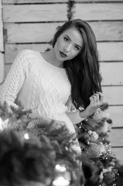 Beautiful sexy woman with Xmas tree in background sitting on elegant chair in cozy scenery. Portrait of girl posing pretty  short tight fit white dress. Attractive brunette female, indoor shot. — Stok fotoğraf