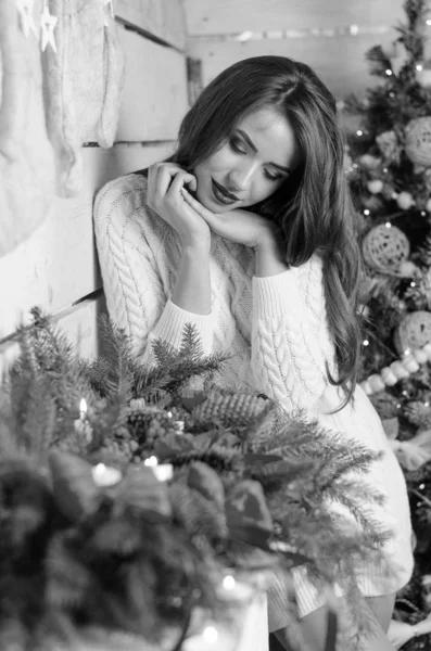 Beautiful sexy woman with Xmas tree in background sitting on elegant chair in cozy scenery. Portrait of girl posing pretty  short tight fit white dress. Attractive brunette female, indoor shot. — Stok fotoğraf