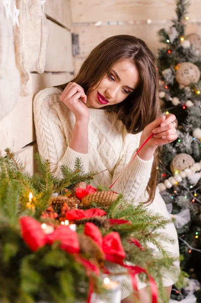 Beautiful sexy woman with Xmas tree in background sitting on elegant chair in cozy scenery. Portrait of girl posing pretty short tight fit white dress. Attractive brunette female, indoor shot. — Stock Photo, Image