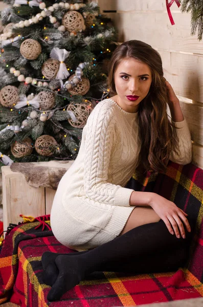 Hermosa mujer sexy con árbol de Navidad en el fondo sentado en una silla elegante en un paisaje acogedor. Retrato de niña posando vestido blanco ajustado bastante corto. Atractiva morena femenina, tiro de interior . —  Fotos de Stock