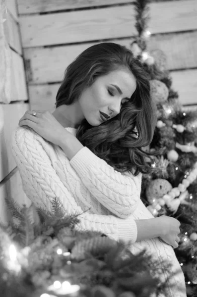 Beautiful sexy woman with Xmas tree in background sitting on elegant chair in cozy scenery. Portrait of girl posing pretty  short tight fit white dress. Attractive brunette female, indoor shot. — 스톡 사진