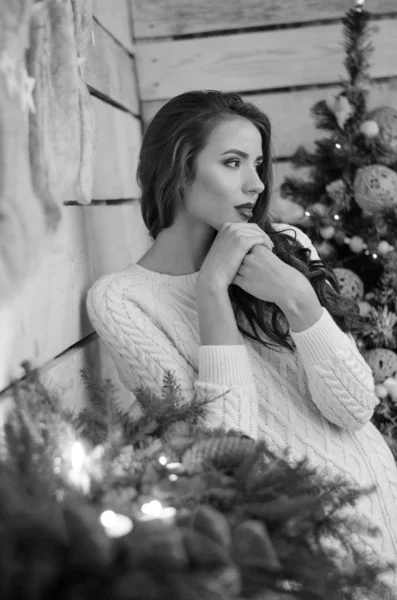 Beautiful sexy woman with Xmas tree in background sitting on elegant chair in cozy scenery. Portrait of girl posing pretty  short tight fit white dress. Attractive brunette female, indoor shot. — Stok fotoğraf