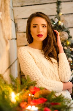 Beautiful sexy woman with Xmas tree in background sitting on elegant chair in cozy scenery. Portrait of girl posing pretty short tight fit white dress. Attractive brunette female, indoor shot.