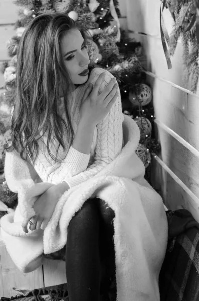 Bella donna sexy con albero di Natale sullo sfondo seduto su una sedia elegante in uno scenario accogliente. Ritratto di ragazza in posa piuttosto corto abito bianco aderente. Attraente bruna femminile, indoor shot . — Foto Stock