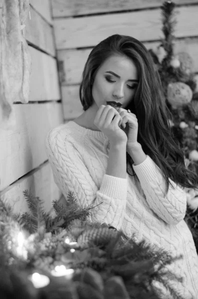 Beautiful sexy woman with Xmas tree in background sitting on elegant chair in cozy scenery. Portrait of girl posing pretty  short tight fit white dress. Attractive brunette female, indoor shot. — Stok fotoğraf