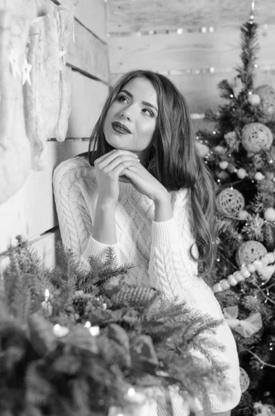 Beautiful sexy woman with Xmas tree in background sitting on elegant chair in cozy scenery. Portrait of girl posing pretty  short tight fit white dress. Attractive brunette female, indoor shot. — Stok fotoğraf