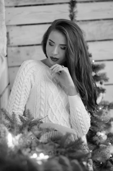 Beautiful sexy woman with Xmas tree in background sitting on elegant chair in cozy scenery. Portrait of girl posing pretty  short tight fit white dress. Attractive brunette female, indoor shot. — 스톡 사진