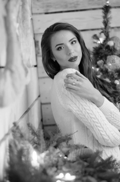 Όμορφη σέξι γυναίκα με χριστουγεννιάτικο δέντρο στο παρασκήνιο κάθεται σε κομψή καρέκλα στο ζεστό τοπίο. Πορτρέτο του κοριτσιού που θέτουν αρκετά σύντομο σφιχτό ταιριάζει λευκό φόρεμα. Ελκυστική μελαχρινή γυναίκα, εσωτερικό πλάνο. — Φωτογραφία Αρχείου