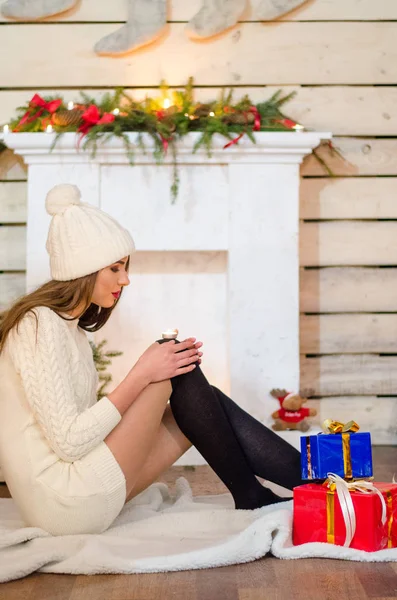 Bella donna sexy con albero di Natale sullo sfondo seduto su una sedia elegante in uno scenario accogliente. Ritratto di ragazza in posa piuttosto corto abito bianco aderente. Attraente bruna femminile, indoor shot . — Foto Stock