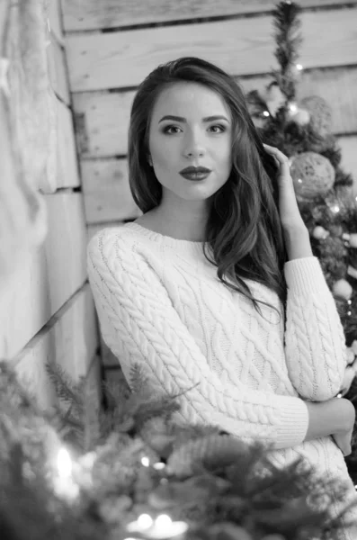 Beautiful sexy woman with Xmas tree in background sitting on elegant chair in cozy scenery. Portrait of girl posing pretty  short tight fit white dress. Attractive brunette female, indoor shot. — Stok fotoğraf