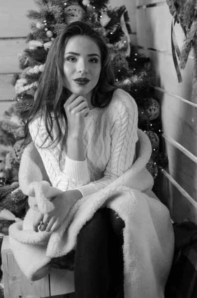 Beautiful sexy woman with Xmas tree in background sitting on elegant chair in cozy scenery. Portrait of girl posing pretty  short tight fit white dress. Attractive brunette female, indoor shot. — Stok fotoğraf