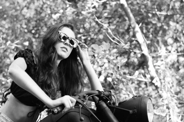 Mujer sexy joven con el pelo ondulado largo en ropa de cuero y gafas de sol posando cerca de una motocicleta. Foto en blanco y negro . — Foto de Stock