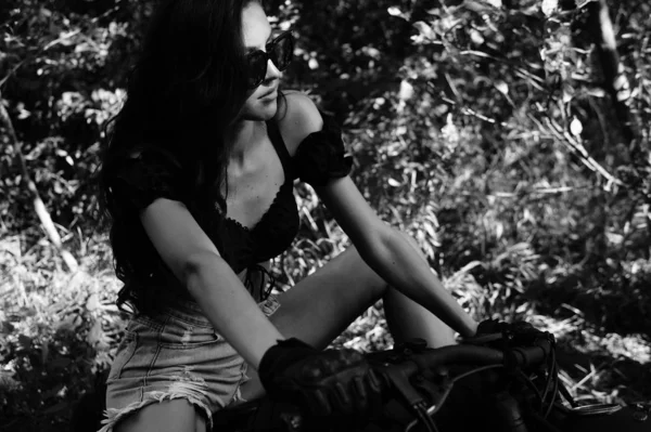 Jeune femme sexy avec de longs cheveux ondulés dans des vêtements en cuir et des lunettes de soleil posant près d'une moto. Photo noir et blanc. — Photo