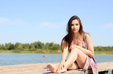 Gölün oradaki tahta iskelede poz veren siyah saçlı kadın. Sırtı Neusiedl Gölü 'nün tahta yaya köprüsüne dönmüş bir kadın. Arka plandaki kayıkhanede güneş yansımaları.