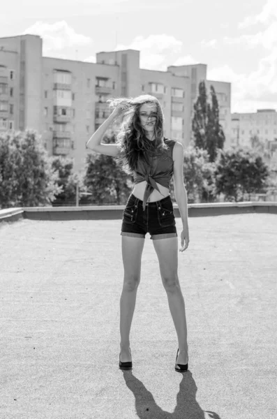 Brünettes asiatisches Mädchen auf dem Dach vor blauem Himmel und modernen Häusern. Junge Frau mit fliegenden Haaren trägt lässiges Kleid - Jeans Shorts schwarzes Hemd — Stockfoto