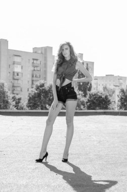 Mavi gökyüzüne ve modern evlere karşı çatıda oturan esmer Asyalı kız. Uçan saçlı genç bir kadın günlük kıyafetler giyer. Kot şort siyah gömlek giyer.