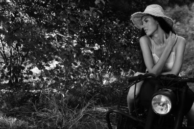 Deri elbiseli, güneş gözlüklü, uzun dalgalı saçlı, motosikletin yanında poz veren genç ve seksi bir kadın. Siyah beyaz fotoğraf.