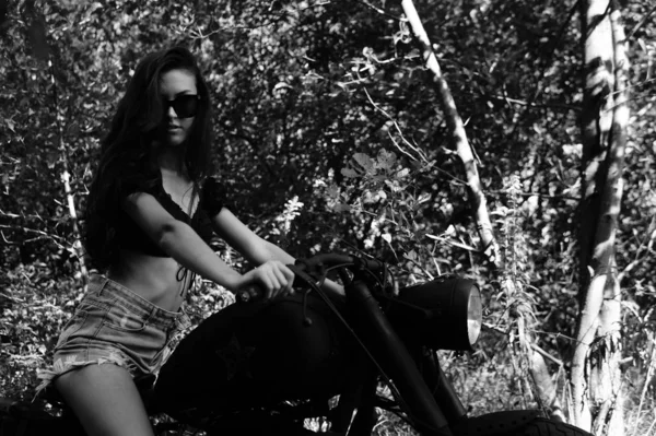 Jovem mulher sexy com cabelos longos ondulados em roupas de couro e óculos de sol posando perto de uma motocicleta. Foto em preto e branco . — Fotografia de Stock