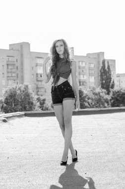 Mavi gökyüzüne ve modern evlere karşı çatıda oturan esmer Asyalı kız. Uçan saçlı genç bir kadın günlük kıyafetler giyer. Kot şort siyah gömlek giyer.